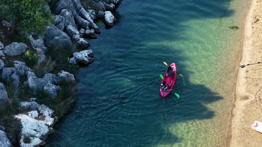 An aerial view of a red kayak with people paddling down a calm, clear river next to a sandy beach and rocky shore.