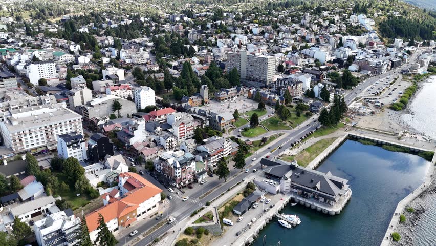 SAN CARLOS DE BARILOCHE, ARGENTINA: High-angle drone photograph capturing a detailed panoramic view of the bustling city center and the famous Centro Civico (Civic Center) square in San Carlos de Bariloche, a premier mountain resort town in Patagonia, Argentina. The image showcases the iconic Swiss and German-inspired alpine architecture, featuring rustic stone and timber buildings, steep gabled roofs, and the historic clock tower arranged around the central cobblestone plaza. Running along the coast of Lake Nahuel Huapi is the main lakeside avenue, bustling with local vehicular traffic, separating the dense urban grid from the scenic waterfront promenade. In the lower right corner, the distinct dark-roofed Puerto San Carlos building sits prominently by the water, overlooking a calm rectangular harbor basin. The town