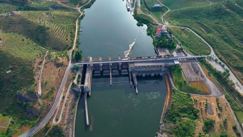 Hydroelectric power station generates electricity using flowing water. Infrastructure includes turbines and dam structures by a river. Surrounding landscape features hills and vegetation.