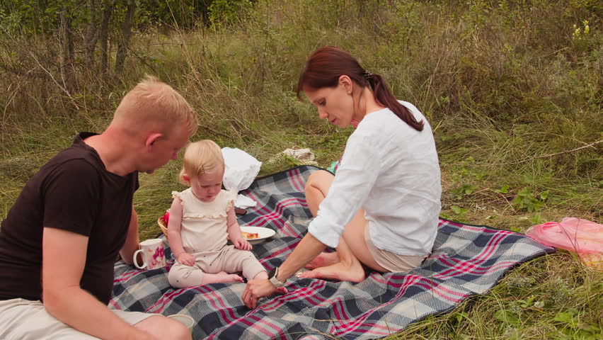 Family with baby sitting on picnic blanket and eating simple food outdoors in countryside