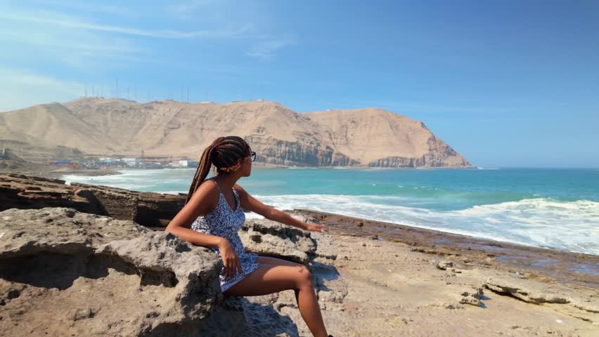 Young african american woman enjoying a sunny day at the beach, sitting on a rock watching the waves crash with mountains in the background