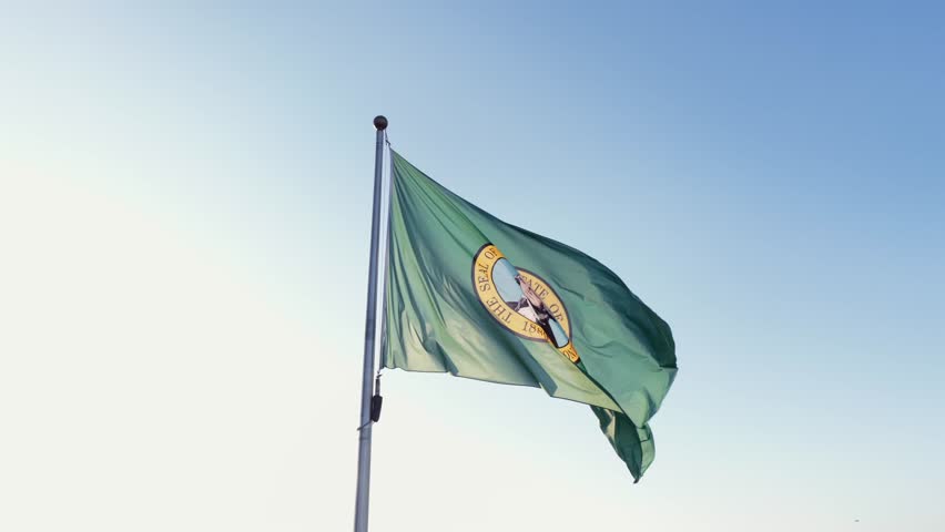 Aerial drone shot of green Washington state flag waving on flagpole against clear blue sky during bright day in Seattle featuring seal of George Washington with sunlight in Washington USA. Close up.
