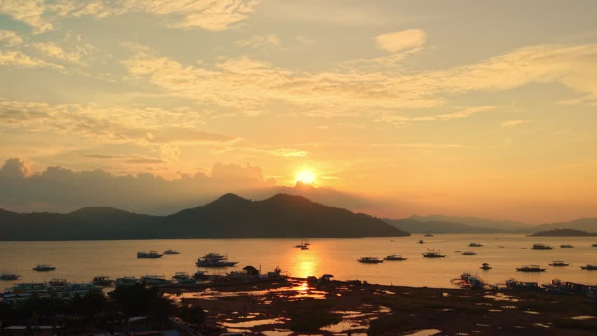 Sunset over a bay in Coron, Palawan, with anchored boats, dark mountain silhouettes, glowing reflection on calm water, layered clouds, and a shoreline foreground.