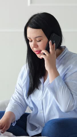 Woman From Asia Speaking Attentively On Phone. Asian Woman Seated On Bed Having Serious Mobile Conversation. An Asian Woman Appears Focused And Serious While Talking On Her Mobile Phone