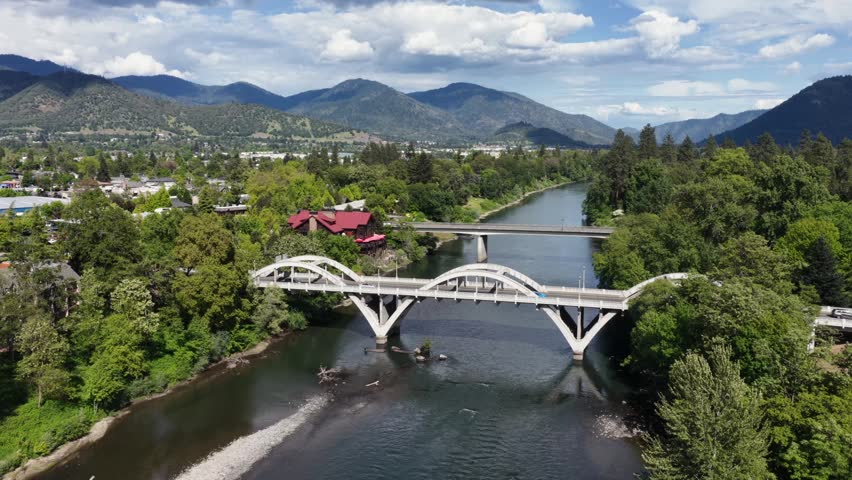 Caveman Bridge spanning Rogue River in Grants Pass Oregon with lush green spring trees and mountain peaks under a sunny sky with clouds in the USA. Aerial wide shot. Sunny day in spring. Approach.