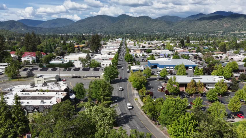 Grants Pass Oregon showing the Caveman Bridge over Rogue River with cars leading into city neighborhood and mountain peaks under a sunny sky in USA. Aerial backwards wide shot. Cloudy day in America.