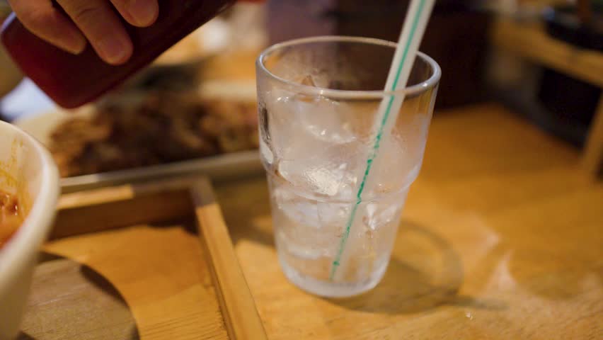 Red hibiscus tea pours into a glass filled with ice cubes on a wooden table