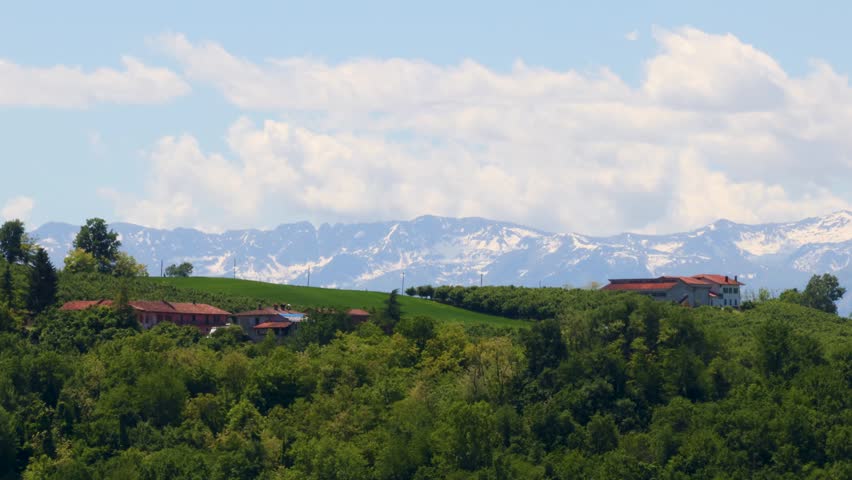 Static wide shot timelapse captures moving clouds above snow-capped mountains and lush green Italian hillsides