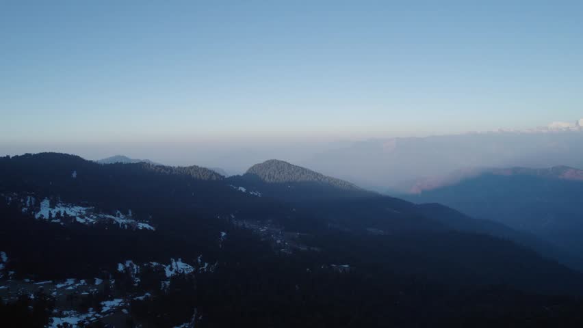 Aerial view of Chopta Tungnath with snow-capped Himalayan peaks and dense evergreen forests under a clear blue sky in Chopta, Uttarakhand, India.