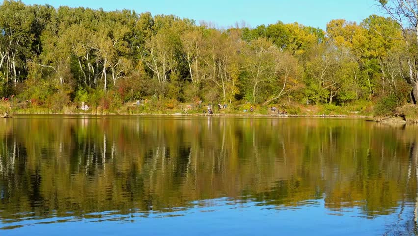 Wide view of lake with tree reflections on rippling water surface. Trees line the far shore while camera remains steady.