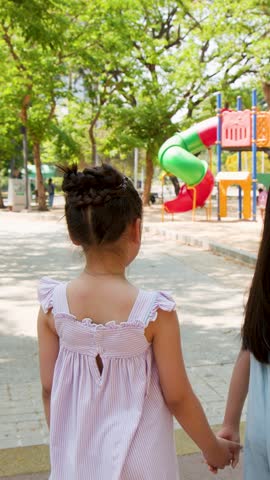 Rear view of two young girls holding hands walking toward a colorful outdoor playground structure