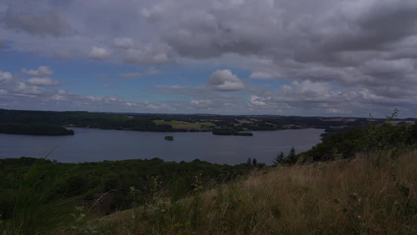 Wide panoramic footage from a hilltop overlooking a large lake with a small island surrounded by dense green forest in Jutland, Denmark. The foreground shows dry summer grass and shrubs on the hillside, while agricultural fields and a small settlement are visible in the distance across the lake under a dramatic partly cloudy summer sky.