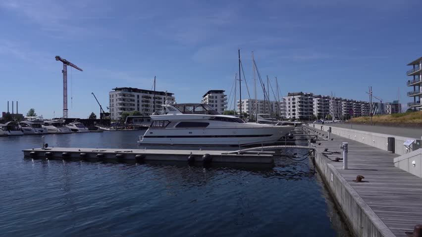 Ground-level footage from the Nordhavn marina in Copenhagen, Denmark. A large white motor yacht is moored at a floating pontoon dock alongside sailboats, with modern white apartment buildings of the newly developed Nordhavn district in the background. A tall red construction crane reflects the area