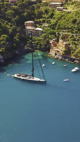 Aerial view of a large sailboat anchored in the turquoise waters of the bay surrounded by lush green hills and colorful villas in Portofino, Liguria, Italy.