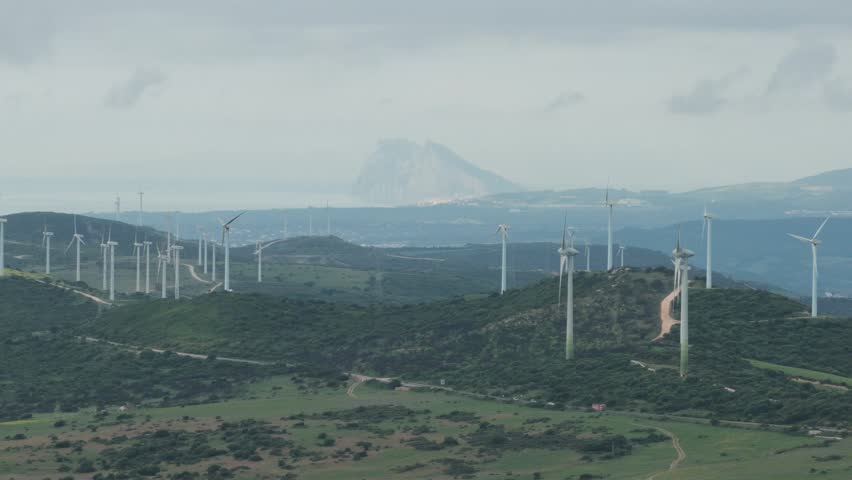 Aerial view of wind turbines on green hills with the Rock of Gibraltar visible in the distance under a cloudy sky Casares, Andalusia, Spain.