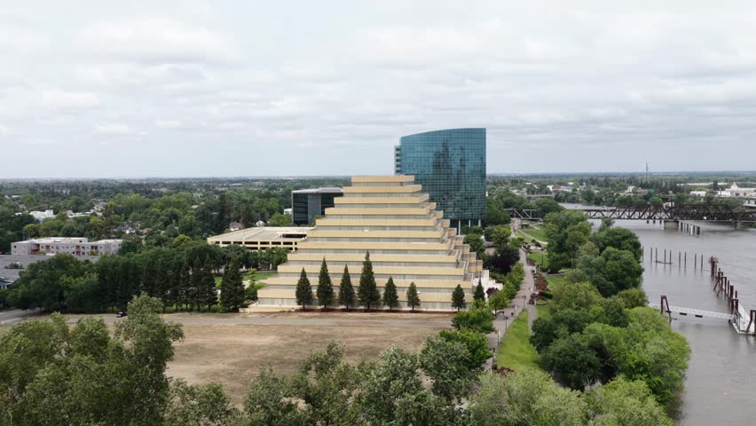 Sacramento Ziggurat Building with stepped pyramid architecture beside modern glass high-rise, Sacramento River waterfront, steel railroad bridge and green trees under cloud sky.Aerial approaching view