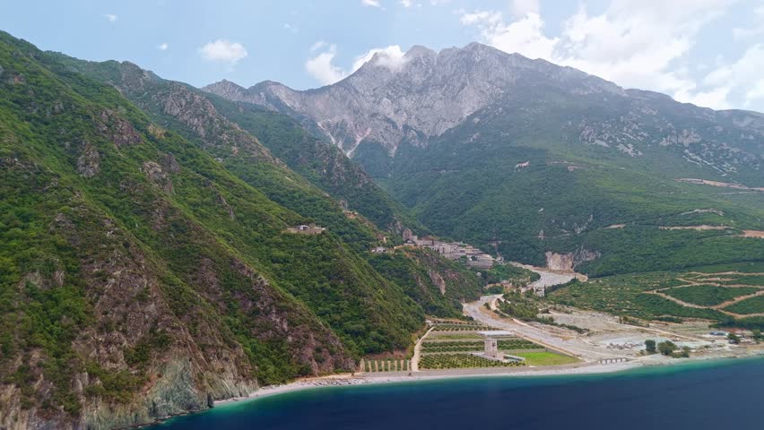 Aerial view of the majestic Holy Athos Mountain rising from the Aegean Sea. Farming fields maintained by monks line the coast beside the turquoise water in the Monastic State of Mount Athos, Greece.