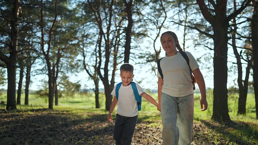 Brother girl hold hands in forest. Trees around happy child sister with backpack. Boy smile walk hike together. Forest path bright sun. Girl walk child brother laugh. Nature trees forest adventure.