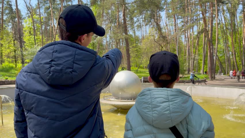 Two brothers in caps looking at park fountain and pointing hands on sunny spring day, back view