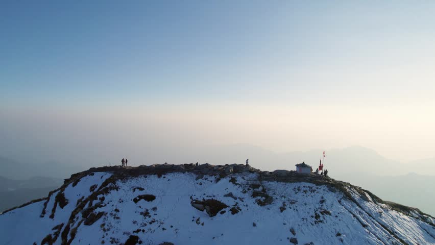Aerial view of Shri Tungnath Temple with people on a snow-covered mountain ridge under a clear sky in Rudraprayag, Uttarakhand, India.