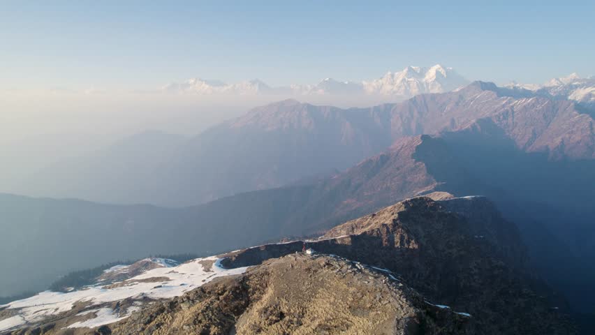 Aerial view of Shri Tungnath Temple nestled on a mountain ridge with snow-capped Himalayan peaks in the distance under a clear sky in Rudraprayag, Uttarakhand, India.