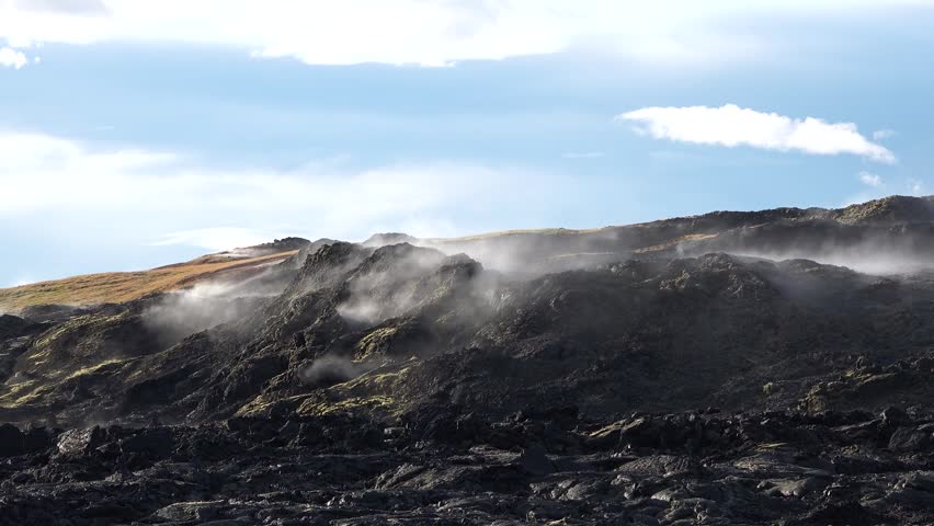 Steam can be seen rising from dark rocks in Iceland. The clouds above create a backdrop as the natural features showcase the unique terrain and volcanic activity in the area.