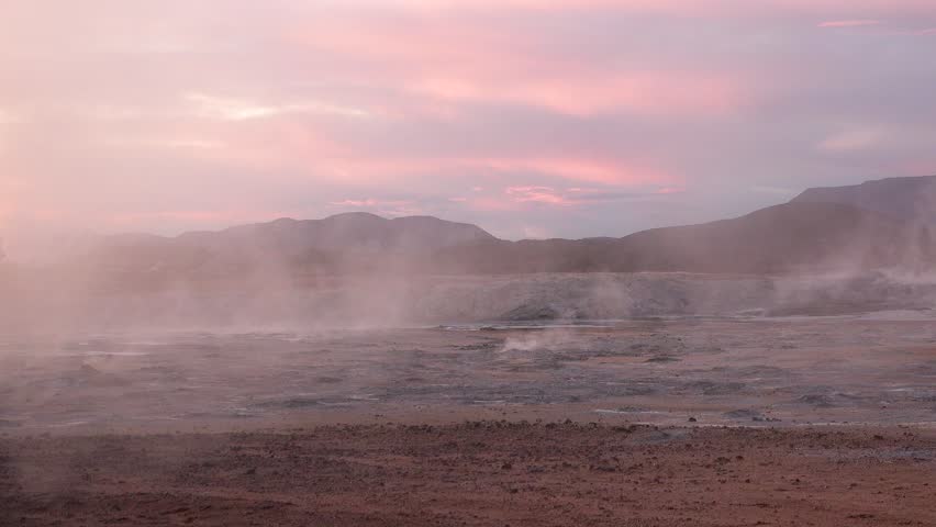 Steam can be seen rising from dark rocks in Iceland. The clouds above create a backdrop as the natural features showcase the unique terrain and volcanic activity in the area.