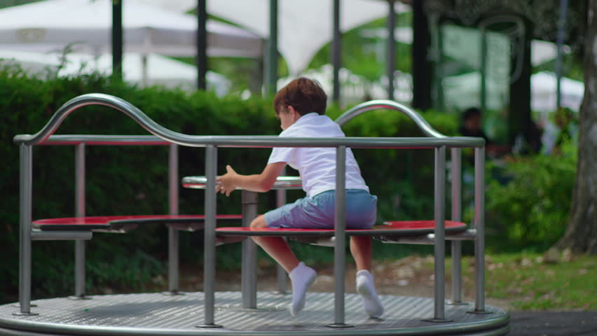 Child spinning merry go round on playground while running along metal rail, lively outdoor childhood scene showing physical activity fun and playful energy
