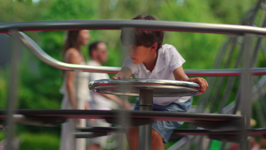 Child riding spinning playground carousel while gripping metal wheel and turning rapidly, lively outdoor scene showing balance coordination and joyful park play