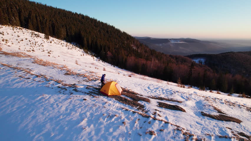 Aerial video shows man near orange tent in snowy Carpathian highlands at sunrise. Atmospheric outdoor scene for trekking, expedition, remote camping, and mountain exploration use