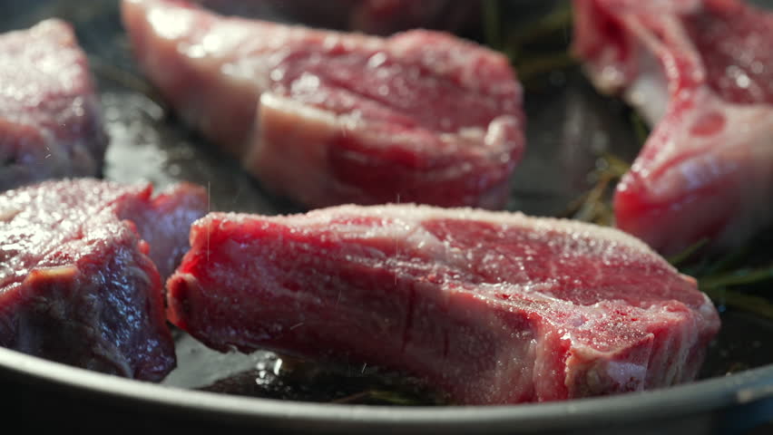 Close up of raw lamb chops in pan with rosemary, showing marbling and fat texture before searing, with natural lighting enhancing surface detail