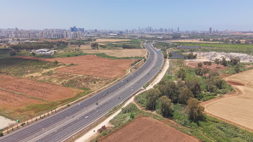 Aerial drone view of a major highway stretching toward Tel Aviv skyline through agricultural fields near Hod Hasharon
