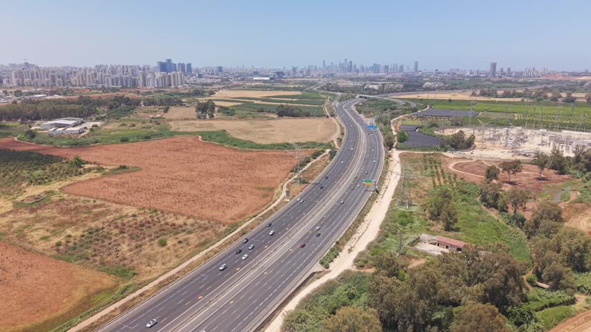 Aerial drone view of a busy multi-lane highway leading toward the Tel Aviv skyline through agricultural fields near Hod Hasharon
