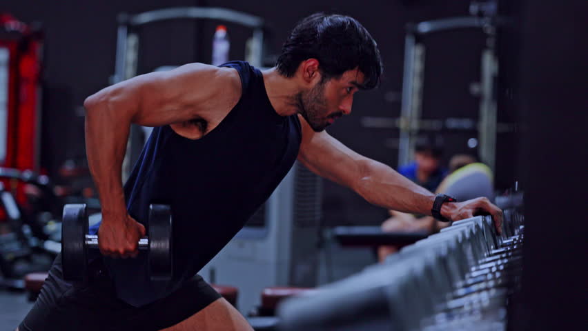 Fit man taking brief rest after intense dumbbell workout in dark gym covered with sweat building strong arm muscle showing deep focus and extreme physical exhaustion during heavy indoor training