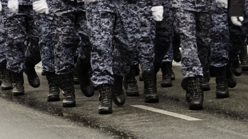 Close-up view of uniformed personnel marching in synchronized formation on a wet street, showcasing black combat boots and digital camouflage trousers during a parade drill