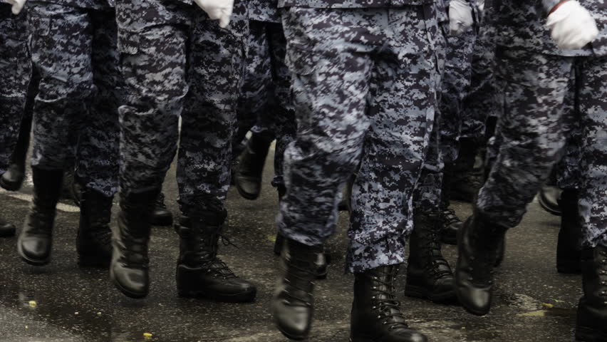 Close-up view of uniformed personnel in blue camouflage marching in formation with black combat boots on a wet street, finishing with a single step as the column passes.