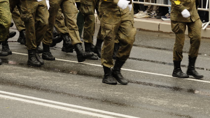 Close-up of synchronized soldiers boots and camouflage trousers marching in tight formation on a rain-soaked city street, with barriers and spectators along the wet roadway