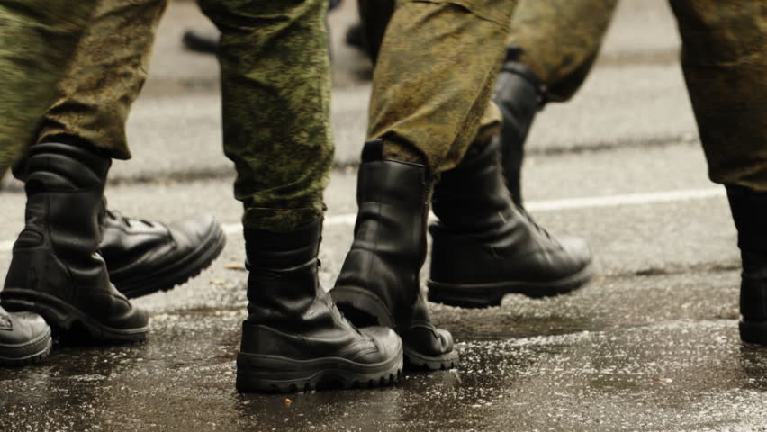 Close-up view of uniformed soldiers in camouflage marching in formation on a wet asphalt road, heavy black combat boots striking the pavement in synchronized steps during drill