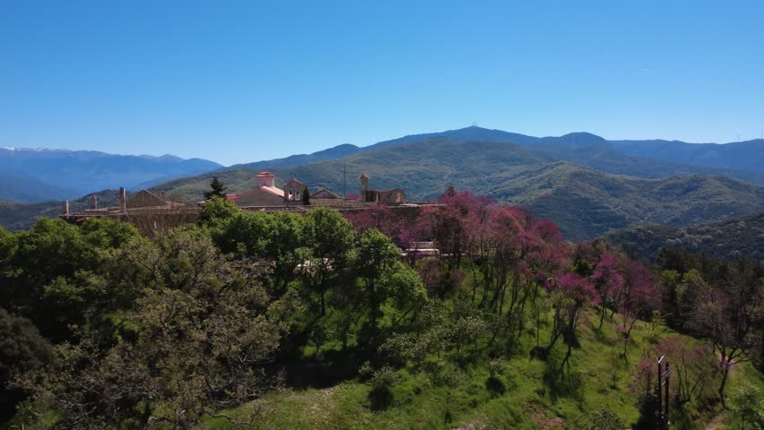 Rotating panoramic establishing shot of a hilltop monastery village surrounded by dense forest and blooming spring trees beneath a clear blue sky in Greece, Europe.