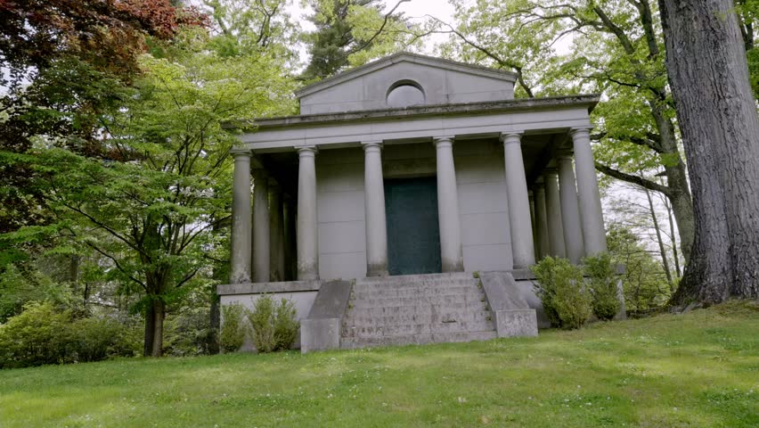 A grand Neo-classical stone mausoleum featuring Doric columns and a triangular pediment sits atop a grassy hill. Surrounded by lush green trees and vibrant spring foliage, this historic burial vault represents timeless architectural beauty in a peaceful graveyard setting.