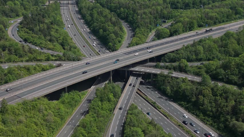 Aerial view of cars and trucks moving on a busy multi-level highway interchange. Vehicles navigate the complex road system nestled among lush green forests on a bright day.