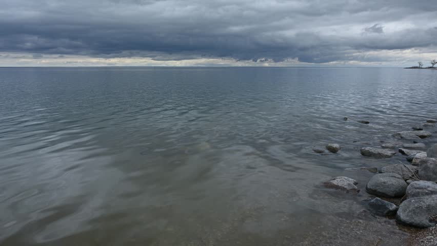 Video of a large lake with rocks along the shoreline. The water has small rippling waves reflecting a cloudy summer sky