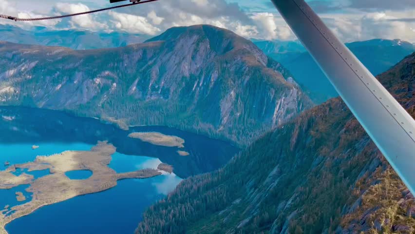 Cinematic aerial view from a floatplane flying over remote Alaskan mountains, turquoise lakes, and wilderness landscape under dramatic clouds.
