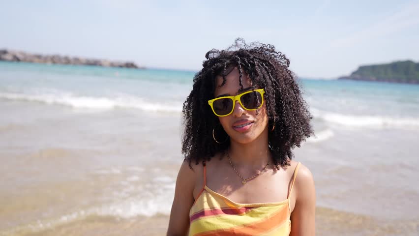 Beautiful mixed race woman with curly hair and yellow sunglasses smiling and drinking a refreshing fruit cocktail from a pineapple shaped cup on a sunny day at the seaside, enjoying her vacation