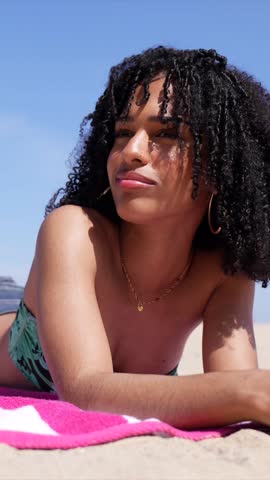 Close up of a beautiful young african american woman with curly hair relaxing on a pink towel, enjoying the warm sun and gentle breeze during her summer vacation on a sandy beach