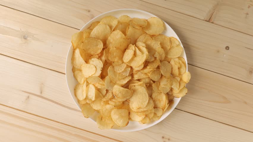 Rotating white plate filled with salty and crispy potato chips on a light wooden background