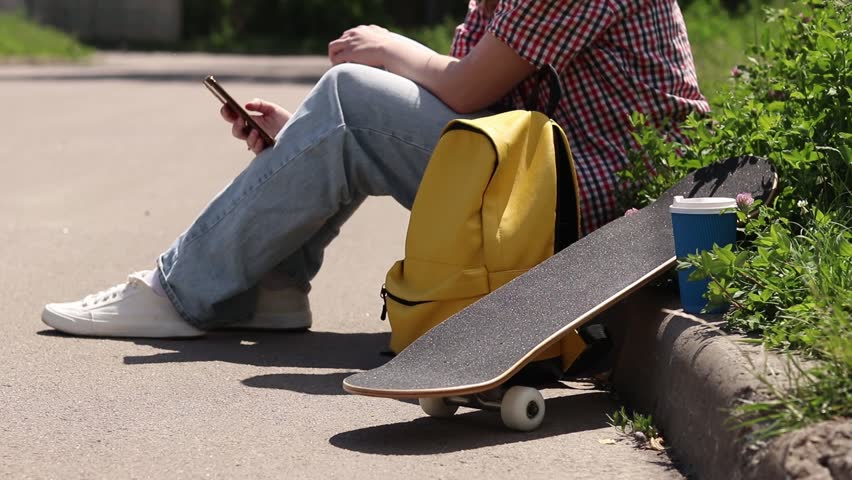 Young woman sitting on the sidewalk with a yellow backpack, skateboard and coffee. Concept of urban recreation and youth culture. Modern city lifestyle, rest after skateboarding. Skater