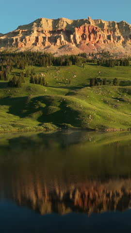 Vertical Screen: Montana, USA scenic landscape featuring dramatic red rock cliffs towering above lush green meadows with conifer forests and pristine alpine lake creating perfect mirror reflections.