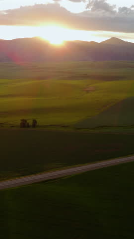 Vertical Screen: Montana, USA aerial drone footage captures stunning golden hour sunset over vast green valley with rolling hills and distant mountain silhouettes along country road.