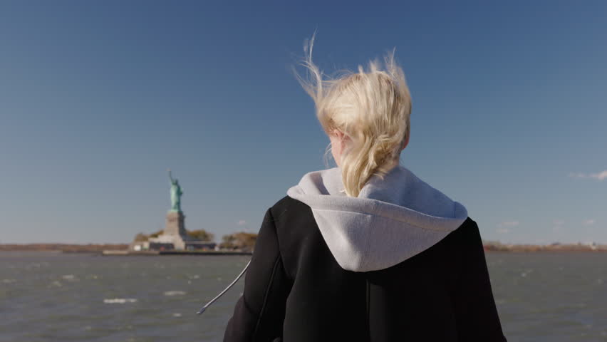 Young woman facing Statue of Liberty on cruise boat in New York Harbor, windy travel scene with iconic landmark, USA.